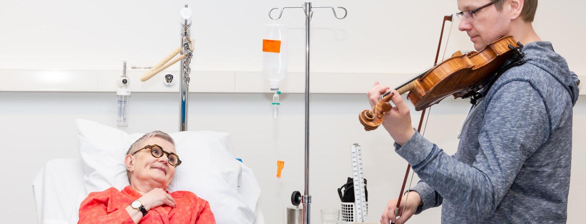 mies soittaa viulua sairaalasängyssä  makaavalle naiselle